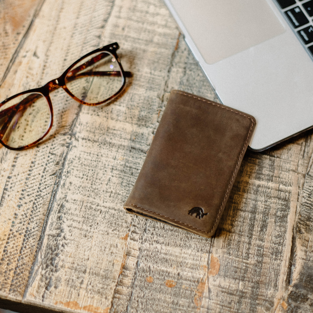The Messenger Wallet in Terra color on a wooden table next to glasses and a laptop