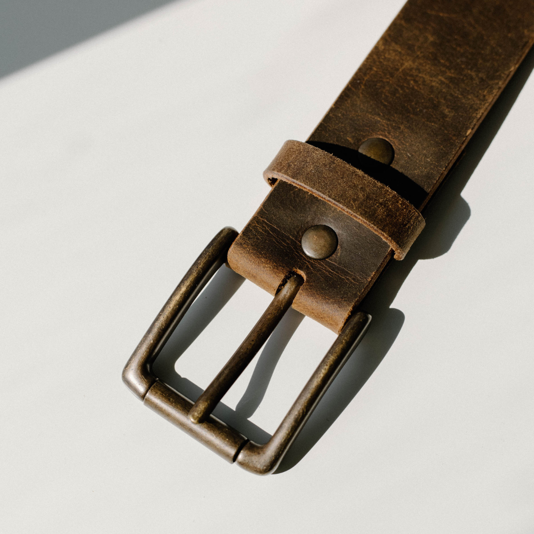 Worn brown leather belt buckle with brass prong and rivet on a light background.