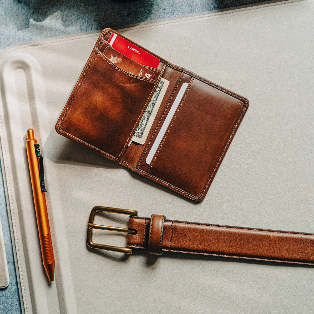 Brown leather wallet with cards and cash, matching leather belt with brass buckle, and orange pen on beige surface.
