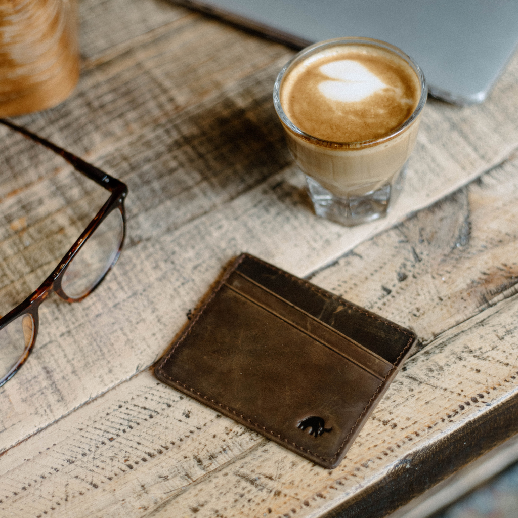 A brown leather card holder on a wooden table next to a coffee cup and glasses.