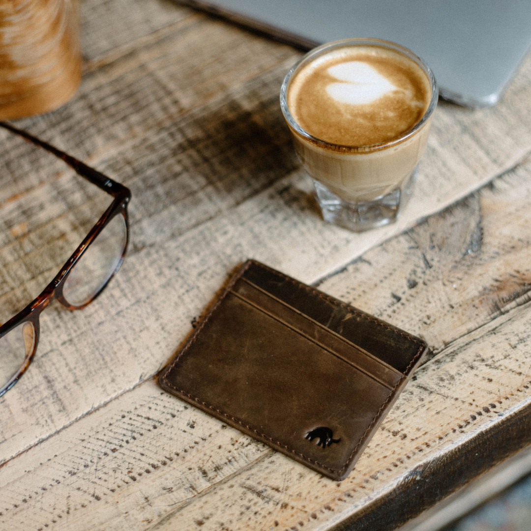A brown leather card holder on a wooden table next to a coffee cup and glasses.