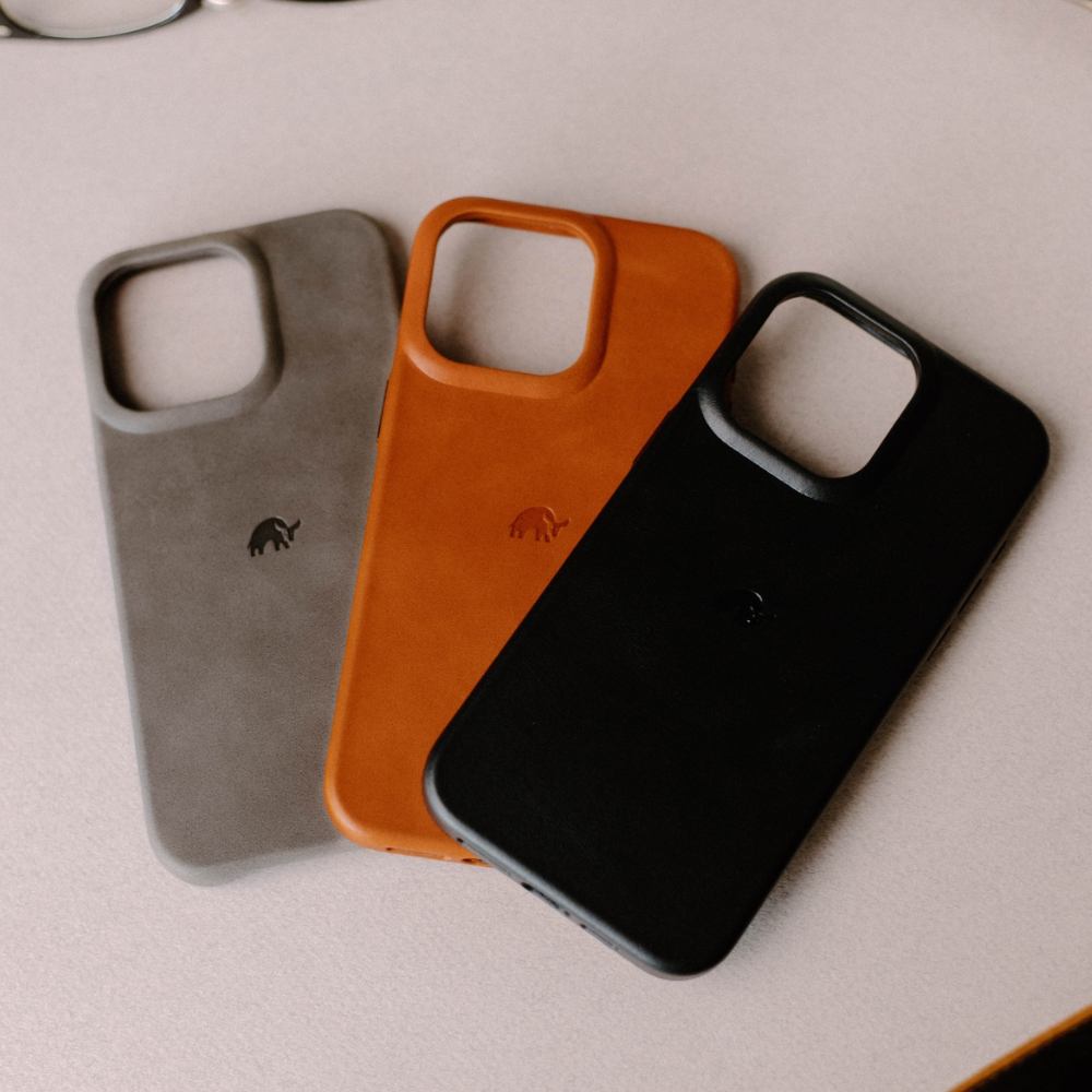 Three leather phone cases (gray, tan, black) fanned on a pale tabletop, embossed small logo on each.