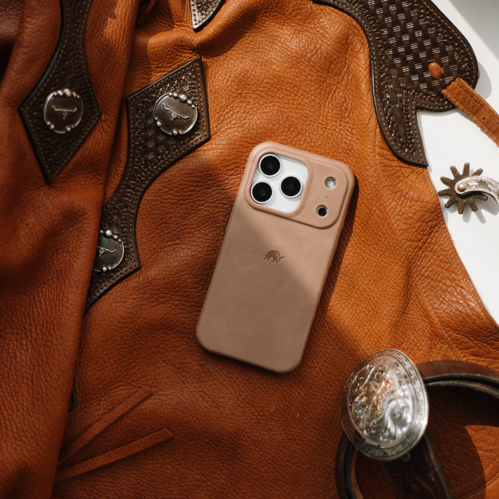 Tan leather iPhone case resting on a brown leather jacket with western silver conchos and leather belt.