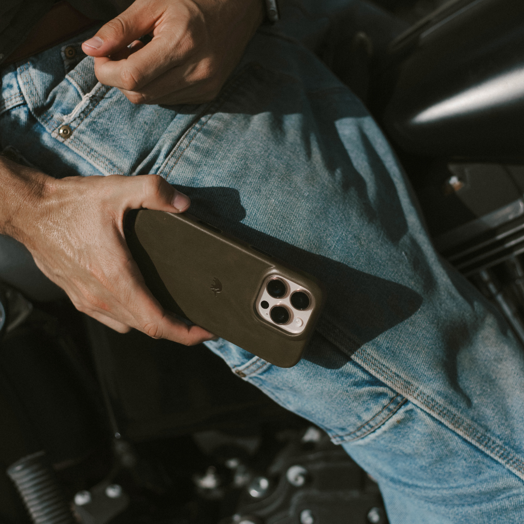 Hand holding a smartphone in an olive-green case, resting on a person's blue-jean thigh with triple camera lenses visible.