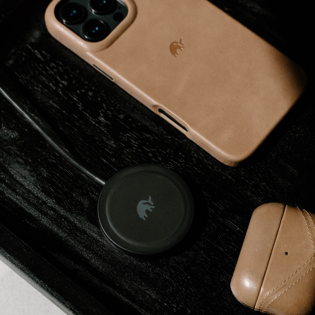 Black wireless MagSafe-style charging puck with cable beside tan iPhone and leather AirPods case