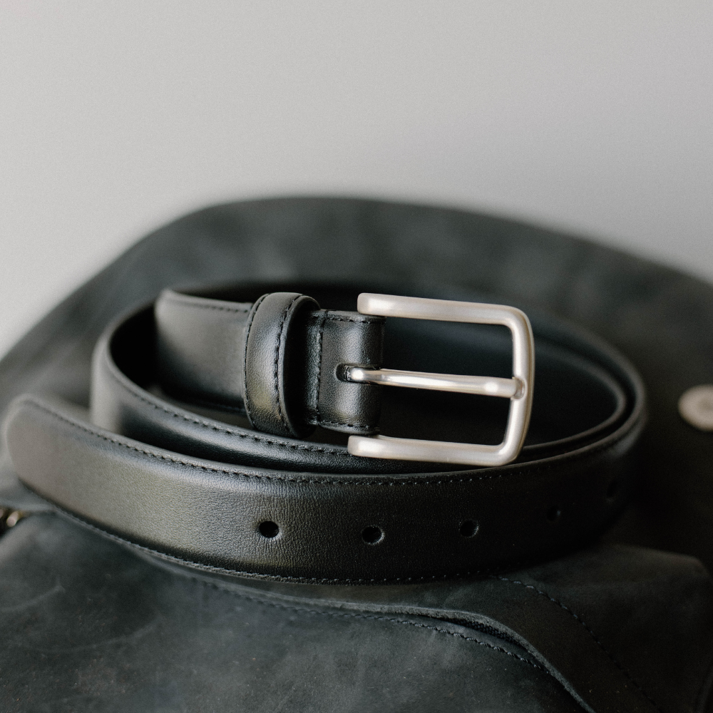 Black leather belt coiled on a dark leather surface, showing a silver rectangular buckle and stitched edge.