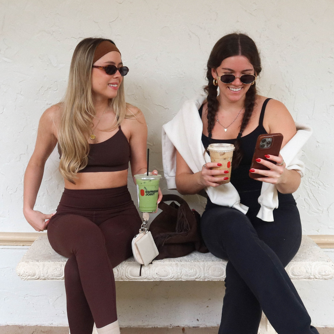 Two women seated on a bench, laughing and holding drinks; a buttercream Carrie wristlet clips to a cup between them