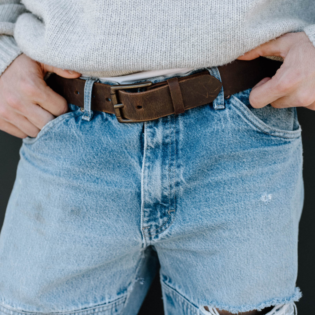 Close-up of person wearing light blue ripped denim shorts and a worn brown leather belt with hands at the waistband.