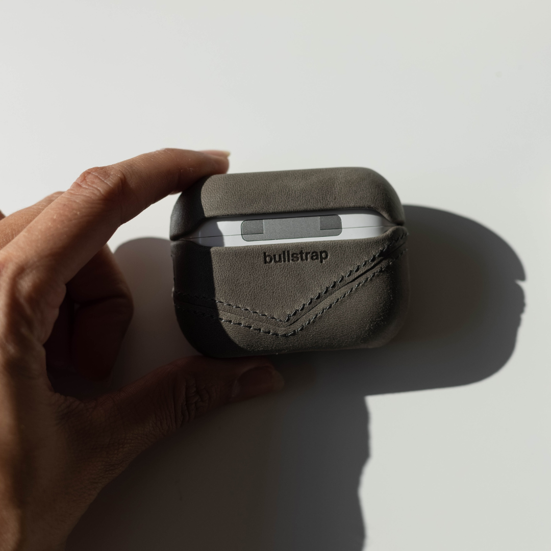 Gray leather AirPods case held in left hand, front embossed bullstrap with diagonal stitching and shadow.
