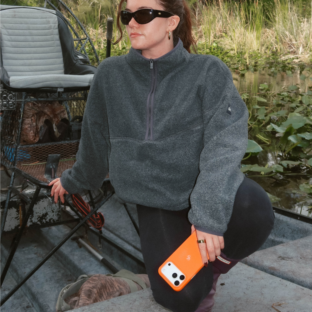Three-quarter view of the Performance Case in Blaze showcasing bright orange leather and camera cutout held by a woman kneeling on a boat