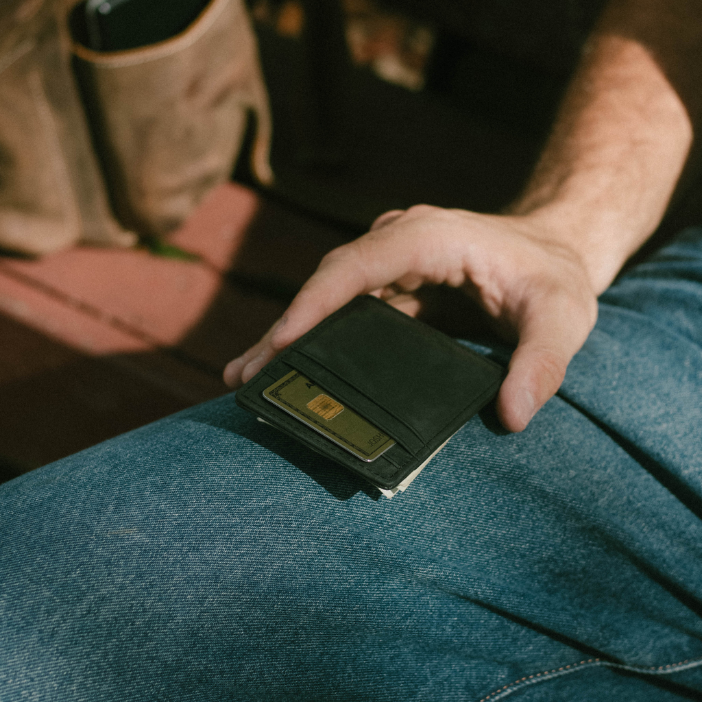 Black leather money-clip wallet on someone's denim-clad lap with a hand reaching toward it