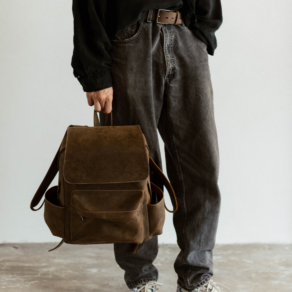 Person's hand holding a brown leather backpack by its top handle; cropped at the waist showing flap and side pockets.