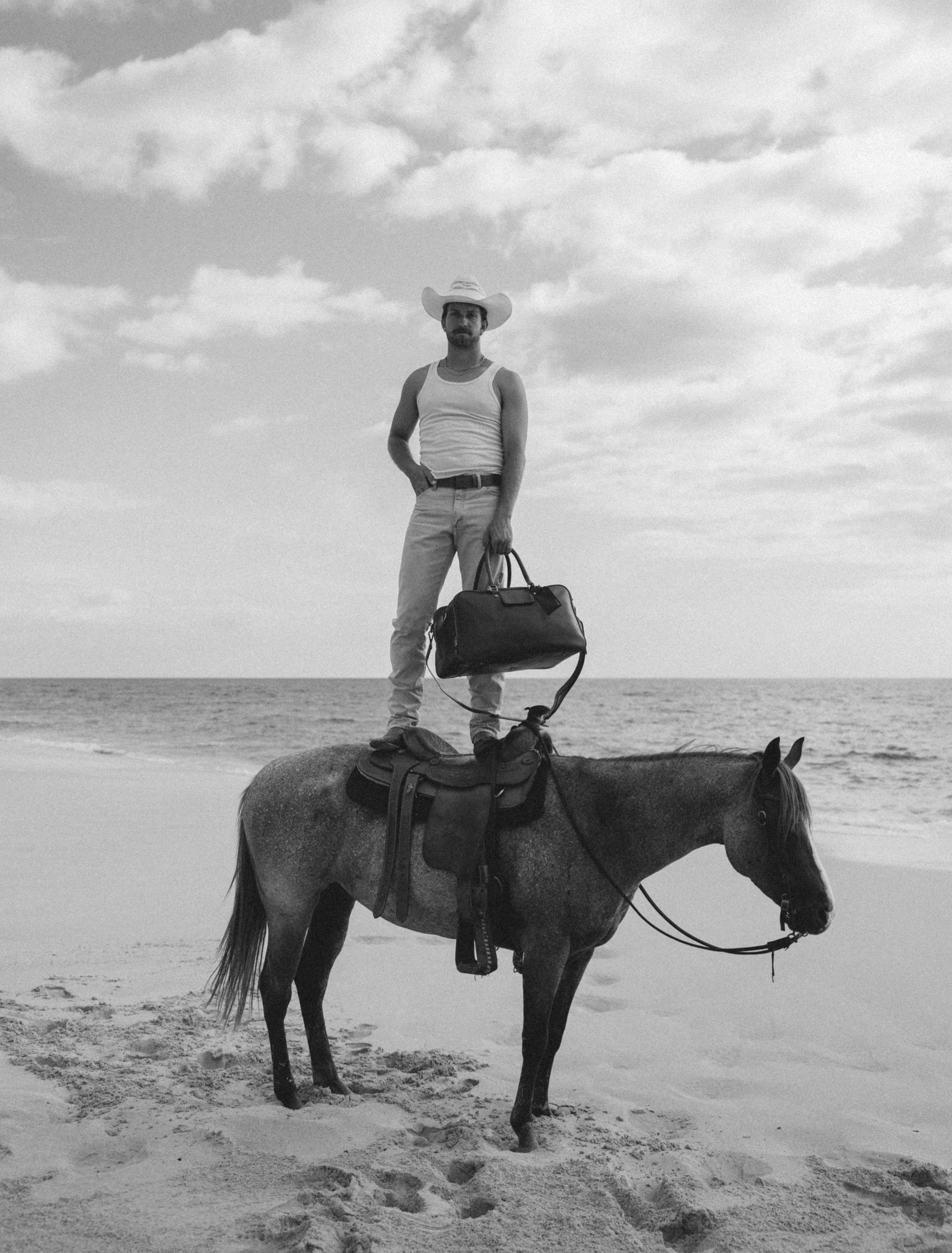 Man in cowboy hat standing on a saddled horse at the beach, holding a leather duffel bag.