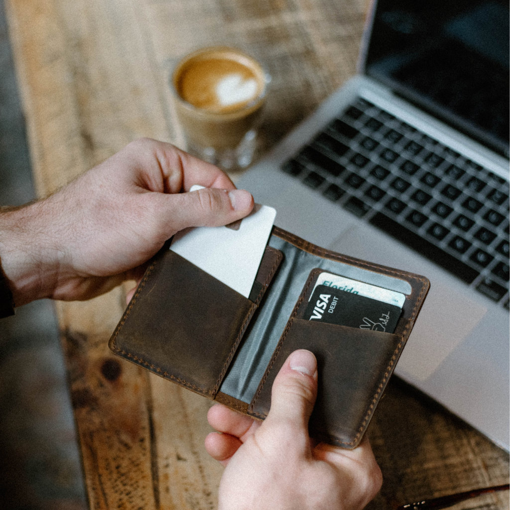 Terra leather messenger wallet open in hands with cards visible; one card reads VISA DEBIT and an ID shows Florida, laptop and coffee behind