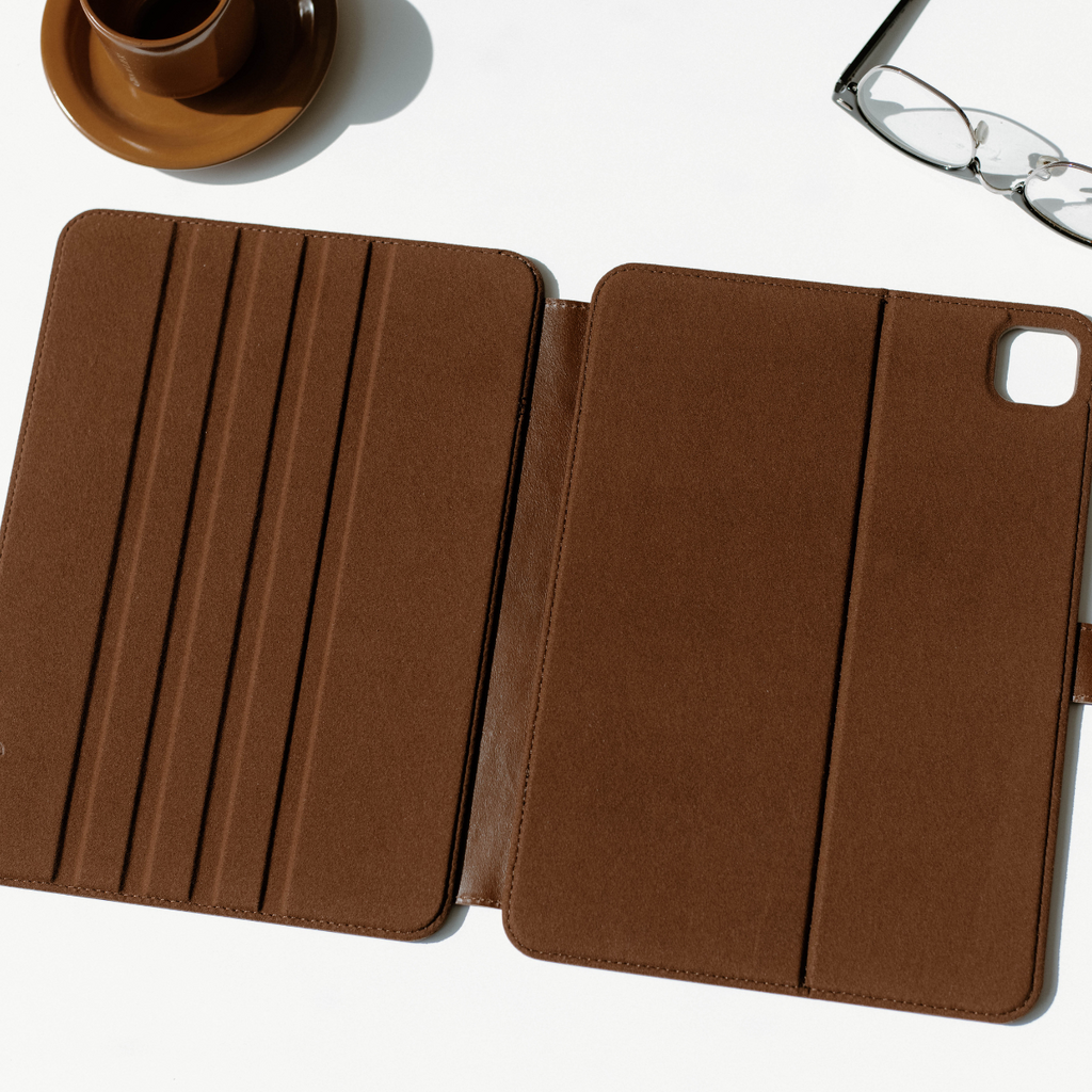 Brown suede folio tablet case opened flat on white surface, with a brown cup at top left and eyeglasses at top right.