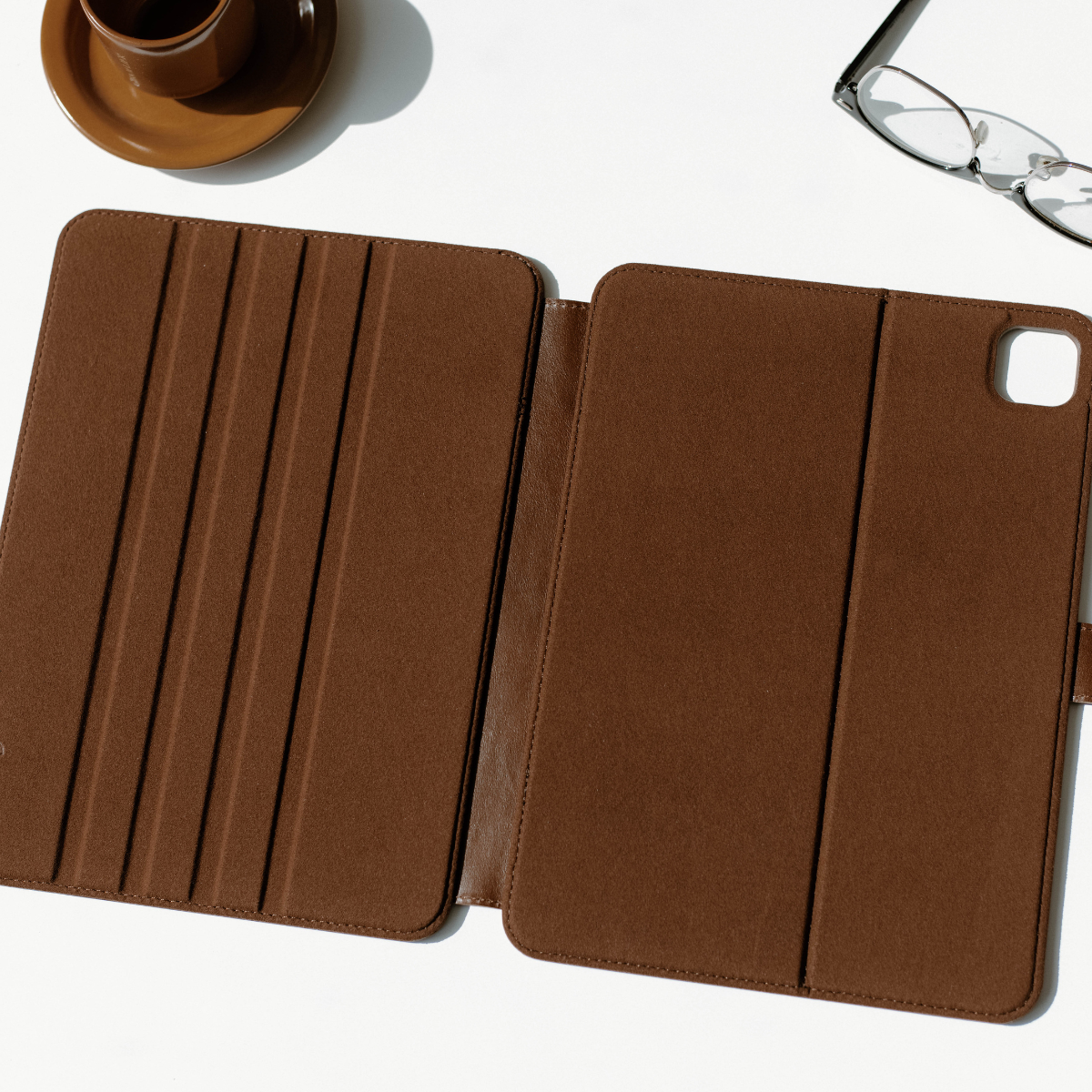 Brown suede folio tablet case opened flat on white surface, with a brown cup at top left and eyeglasses at top right.