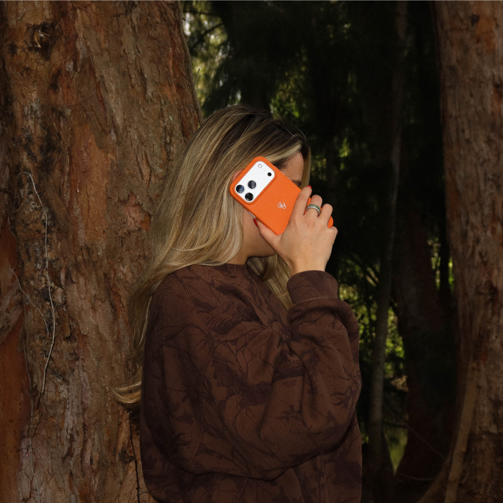 Woman holding iPhone with hunter orange Blaze leather case to her ear in a wooded outdoor setting