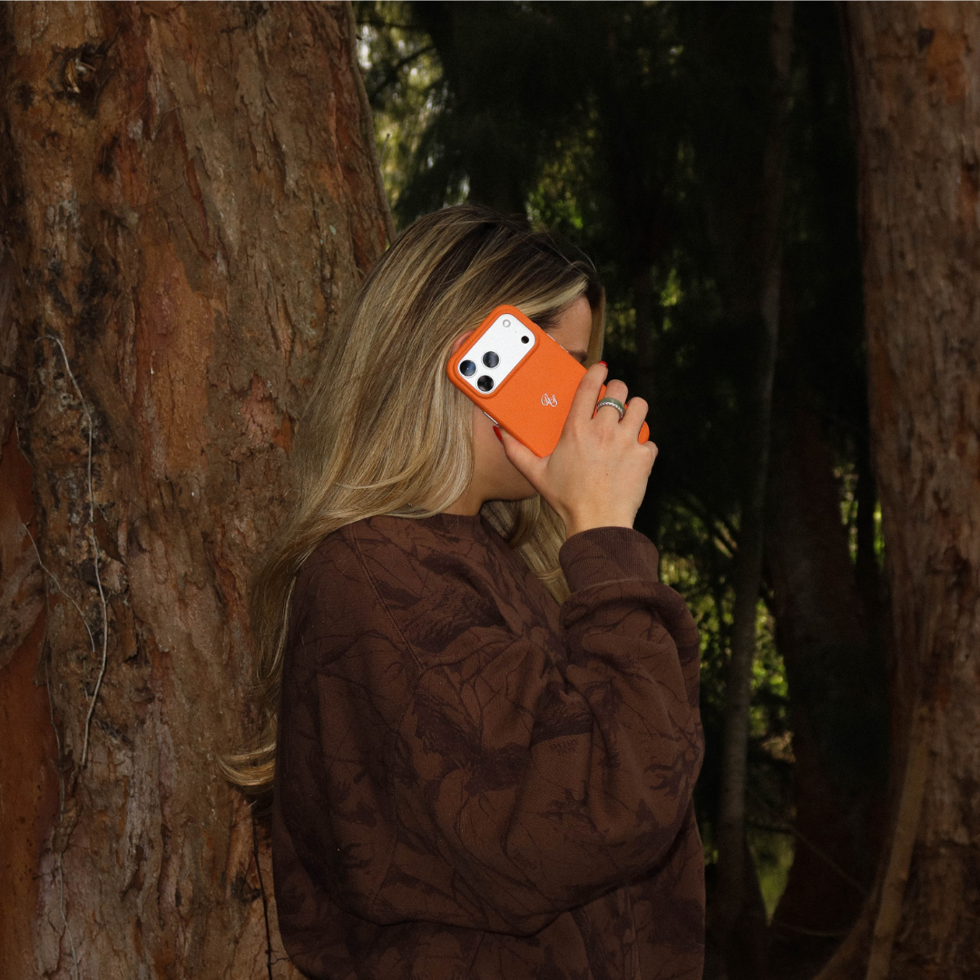 Woman holding iPhone with hunter orange Blaze leather case to her ear in a wooded outdoor setting