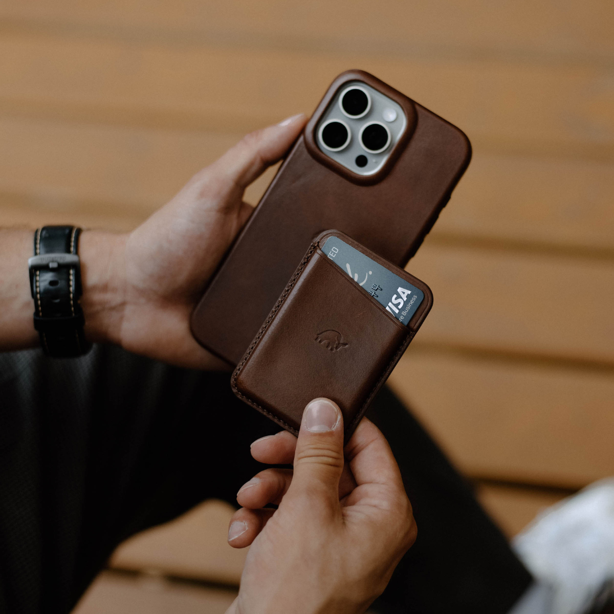 Close-up of hands holding Terra leather Minimalist iPhone case and matching MagSafe wallet with a visible Visa card on a bench
