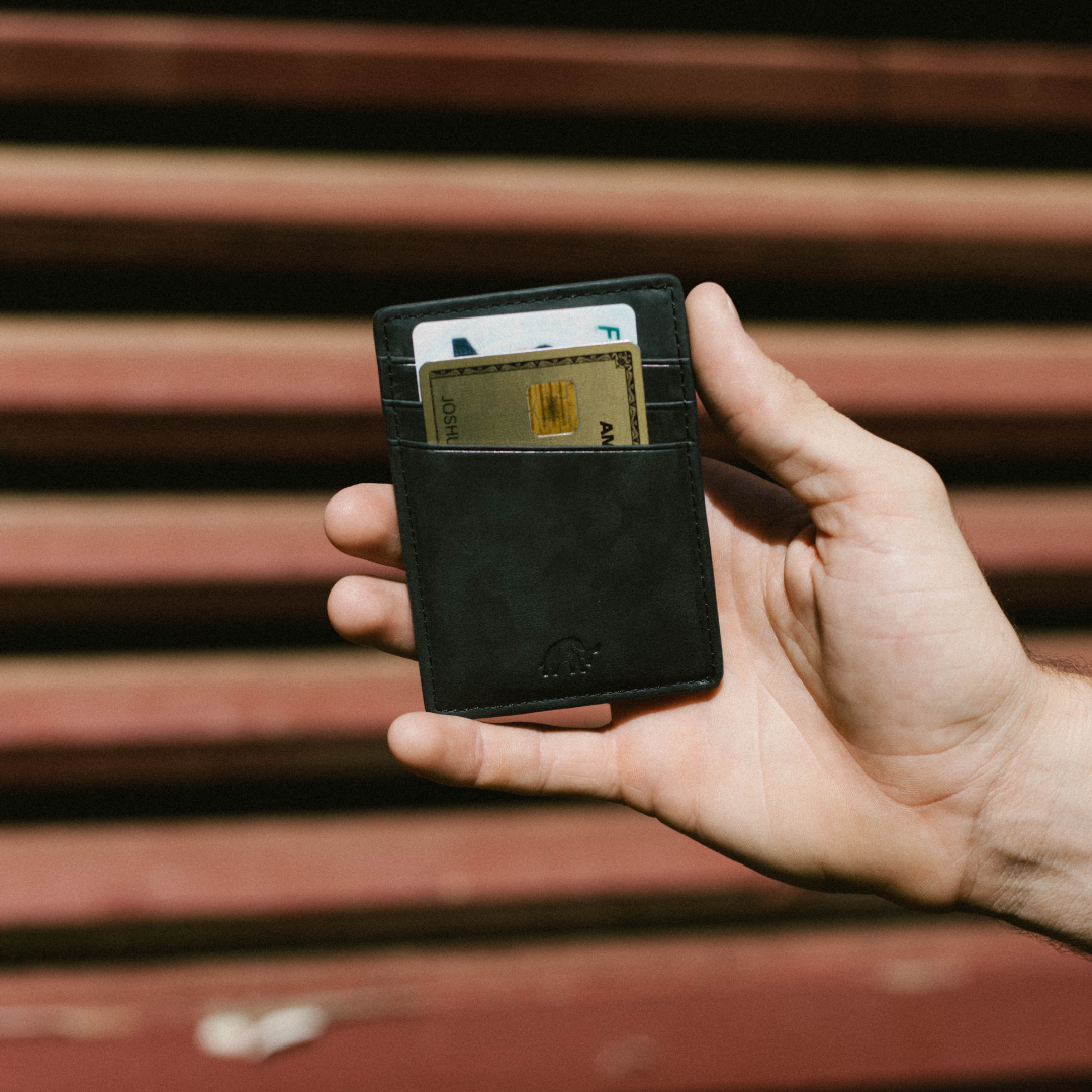 Black slim leather money clip held in a hand, showing a gold chip card labeled JOSH and a white card