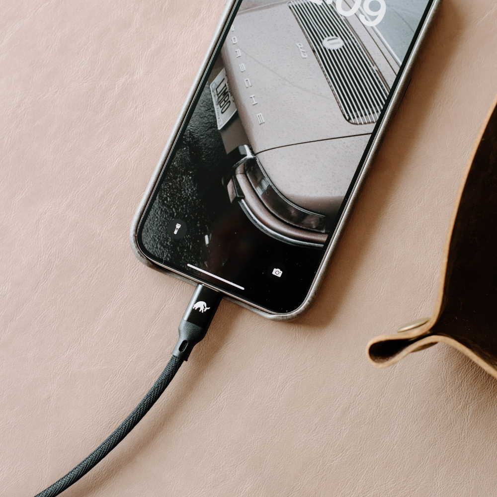Phone showing PORSCHE and 09 on wallpaper, charging via braided cable on tan leather surface