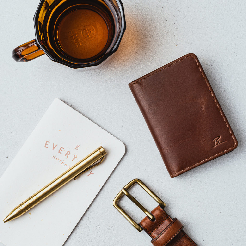 Bullstrap Reserve 'The Minimalist' brown wallet with embossed B beside notebook reading EVERY NOTEBOOK, brass pen, amber cup