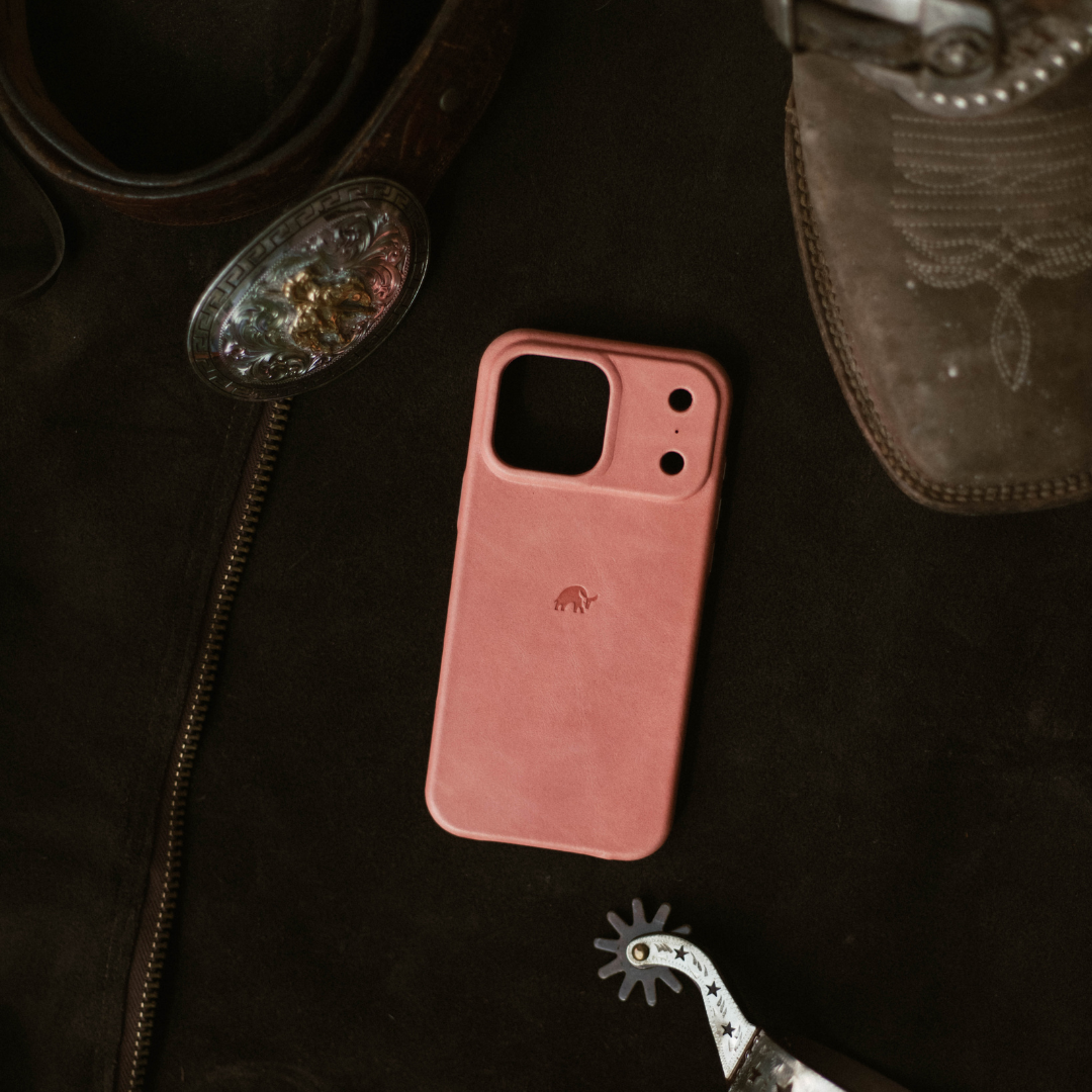 Pink leather minimalist phone case with small elephant logo on dark jacket beside a boot and spur