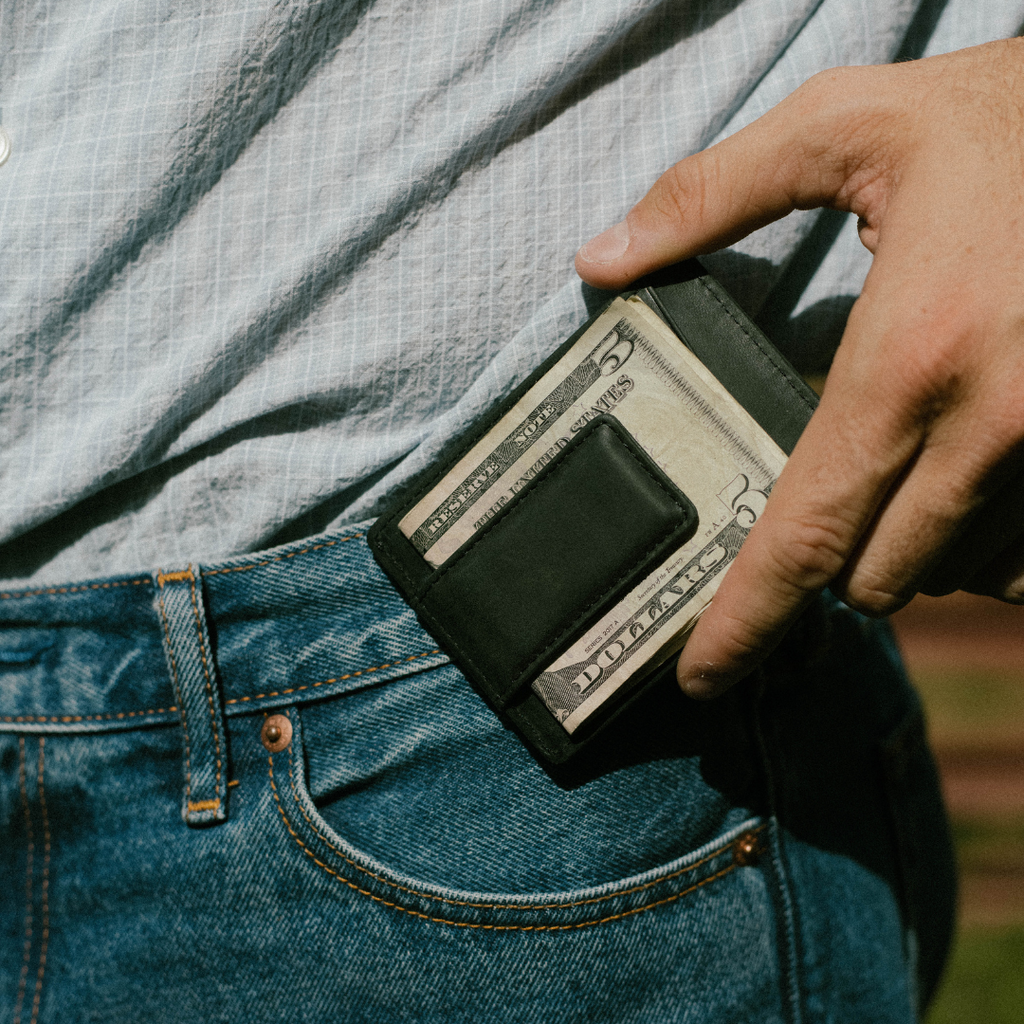 Black money clip holding folded bills showing FIVE DOLLARS UNITED STATES OF AMERICA 5, tucked into blue-jeans front pocket