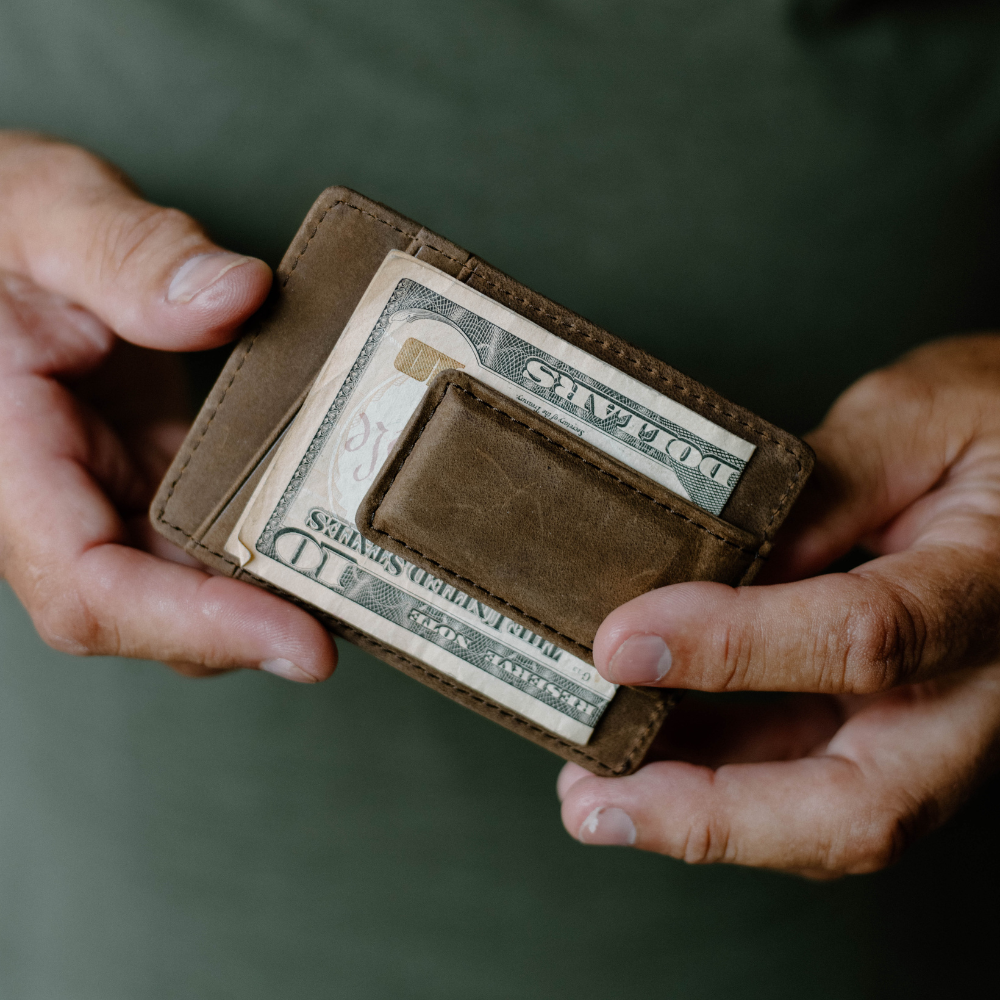 Hands holding a brown money-clip wallet with visible United States DOLLARS folded into the clip.
