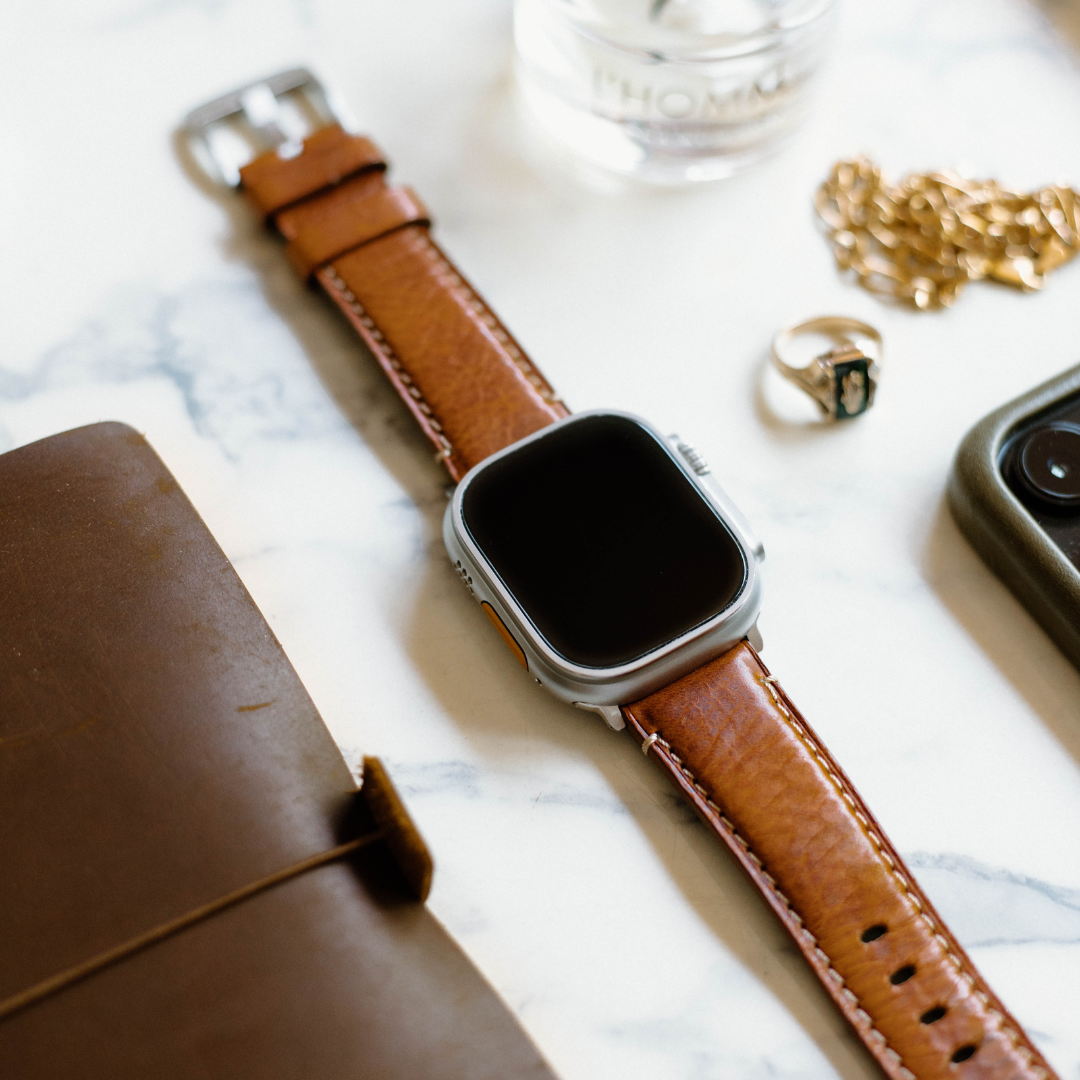 Brown leather Apple Watch strap on a marble surface with accessories