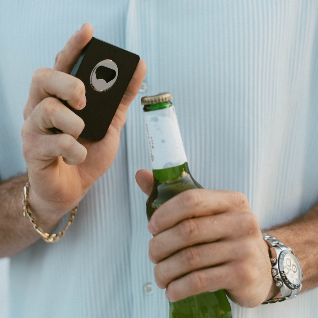 A person holding a black Magnetic Popper Wallet with a built-in bottle opener, ready to open a beer bottle.