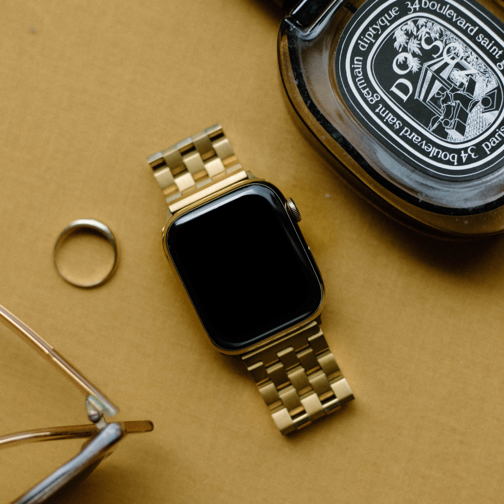 Gold Apple Watch with link strap on tan surface beside coaster labeled 'diptyque 34 boulevard saint germain paris'.