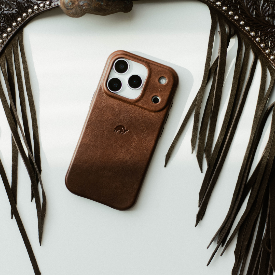 Brown leather iPhone case with raised camera surround and small embossed logo, on white background with dark leather fringe.