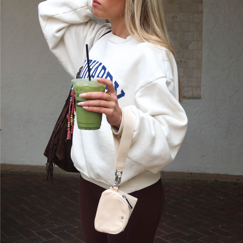 Close-up of The Carrie Wristlet in Buttercream showcasing detachable strap and front zip pocket with model in cream sweatshirt
