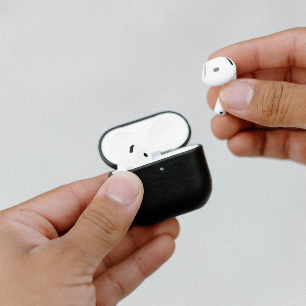 Close-up of black leather AirPods case with lid open showing white AirPods, held in hands against a light gray background