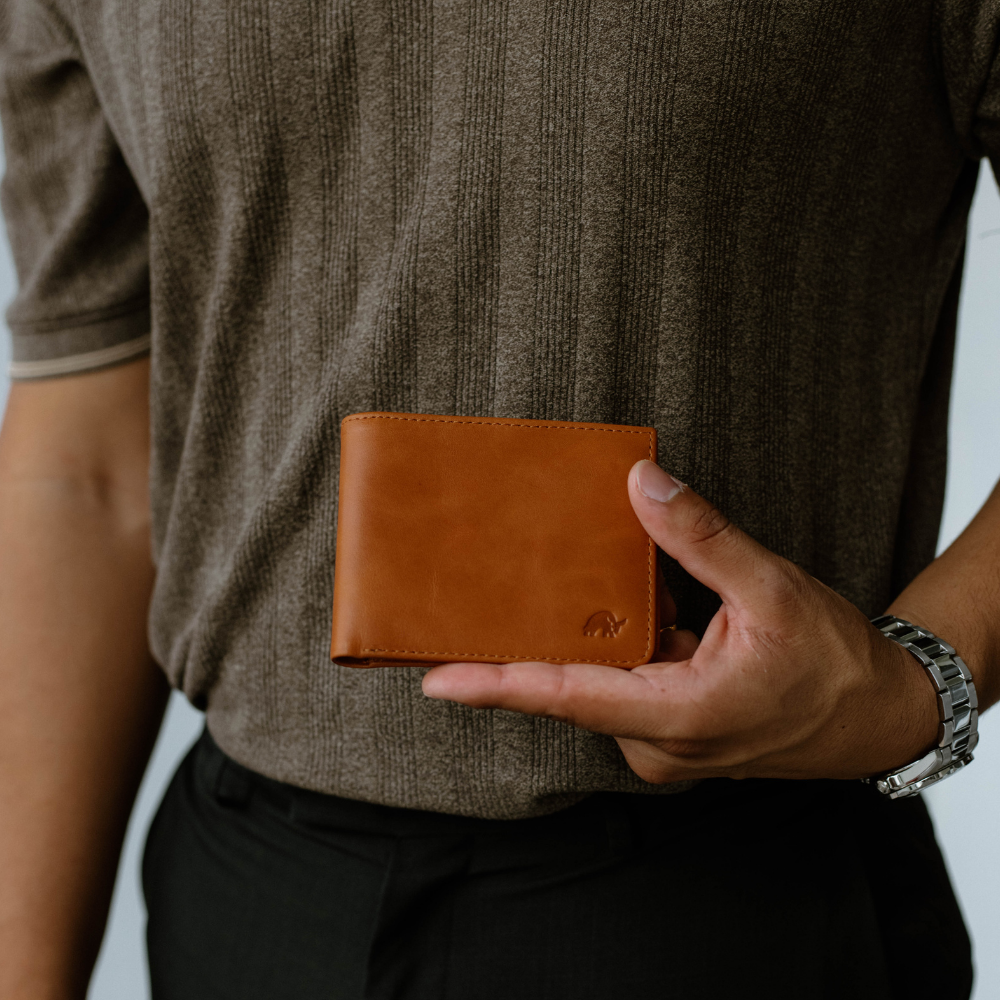 Person holding tan leather bifold wallet at mid-torso against ribbed brown shirt; silver wristwatch visible