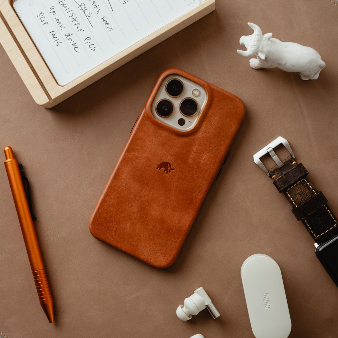 Sienna leather phone case on iPhone with embossed rhino logo, laid on brown surface with pen, earbuds, notebook, and watch