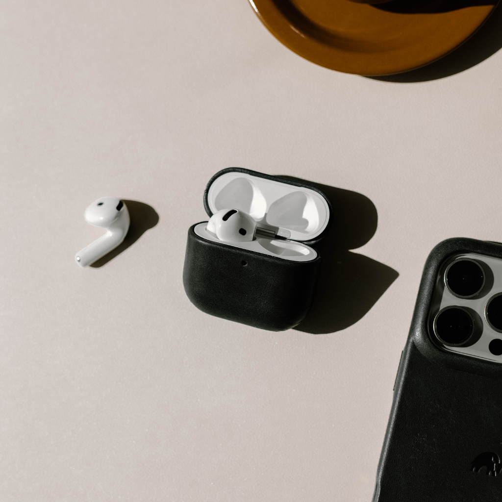 Angled top view of black leather AirPods case open with one earbud inside and one beside it on a pale textured surface