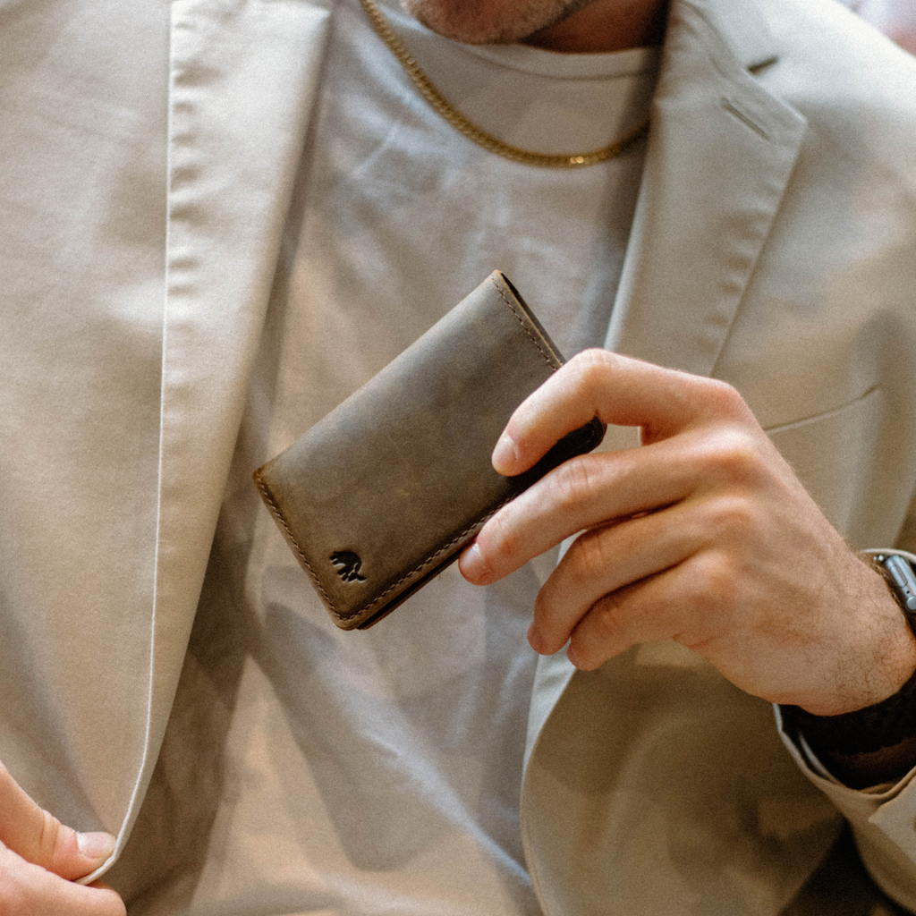 Hand holding the Messenger Wallet - Terra above a beige suit jacket pocket.