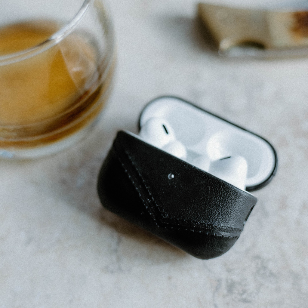 Three-quarter view of black leather AirPods case with stitched front flap holding white AirPods on a light stone surface with blurred coffee cup