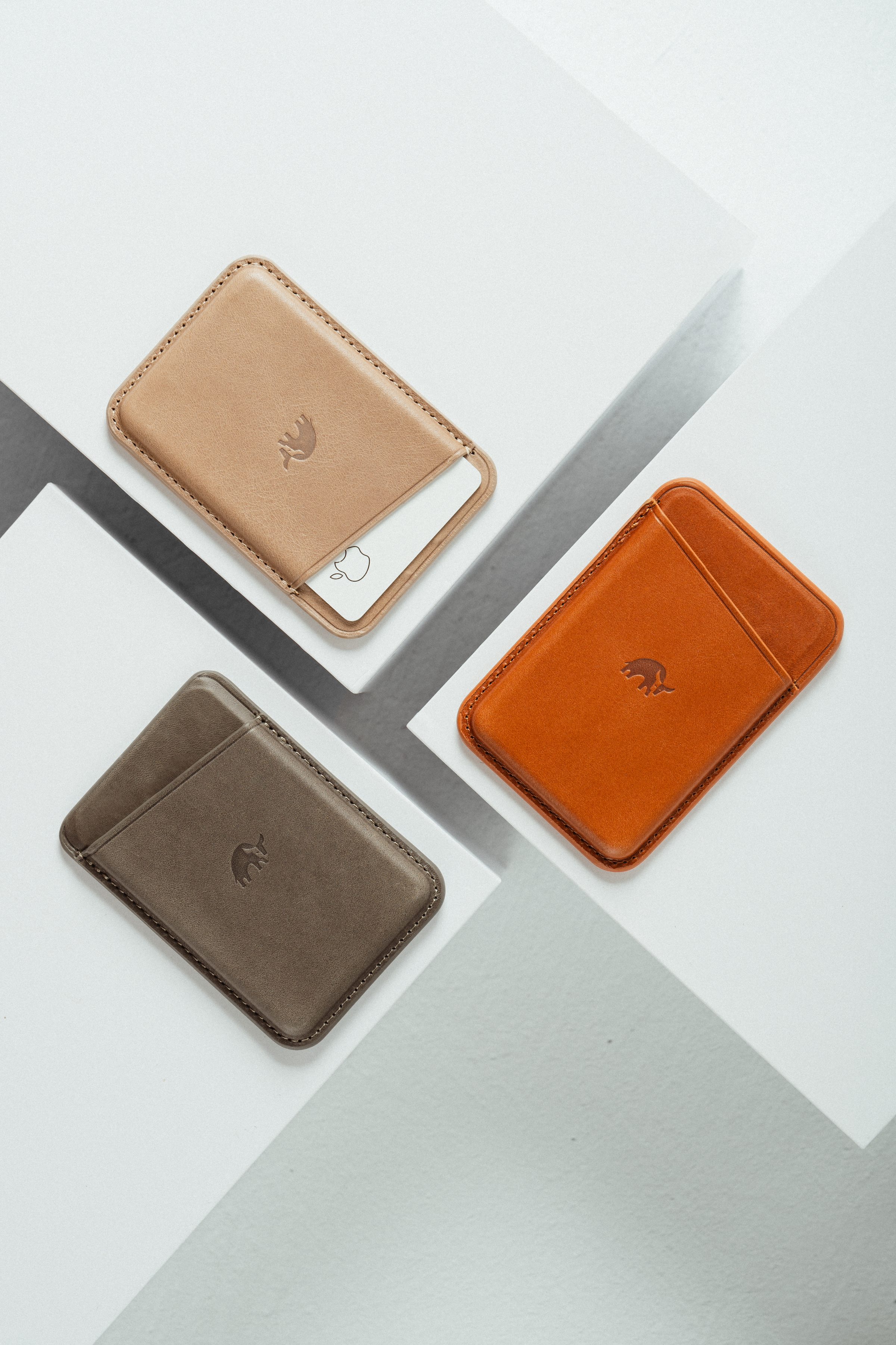 Three slim leather card wallets (tan, gray-brown, orange) on a white geometric surface; tan wallet holds a white card with Apple logo.
