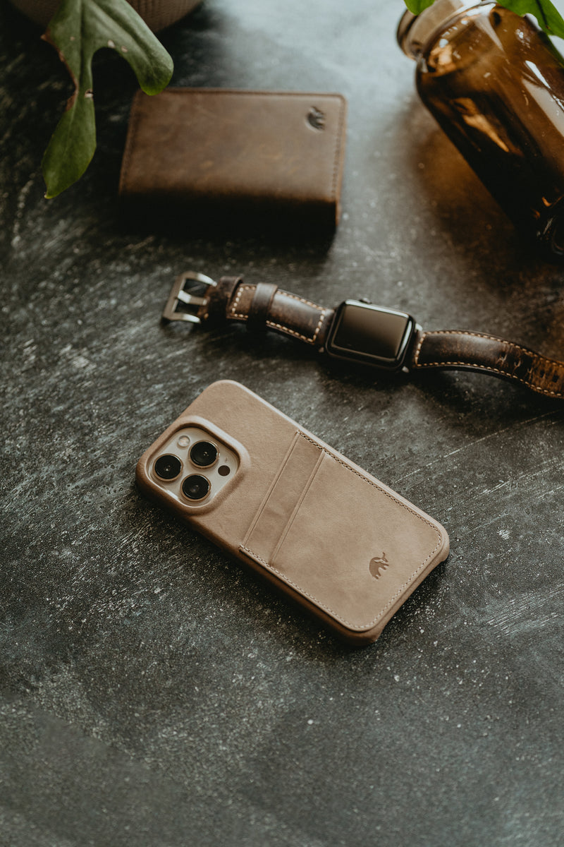A Bullstrap iPhone case in beige color, a brown wallet, and a brown watch on a dark surface.