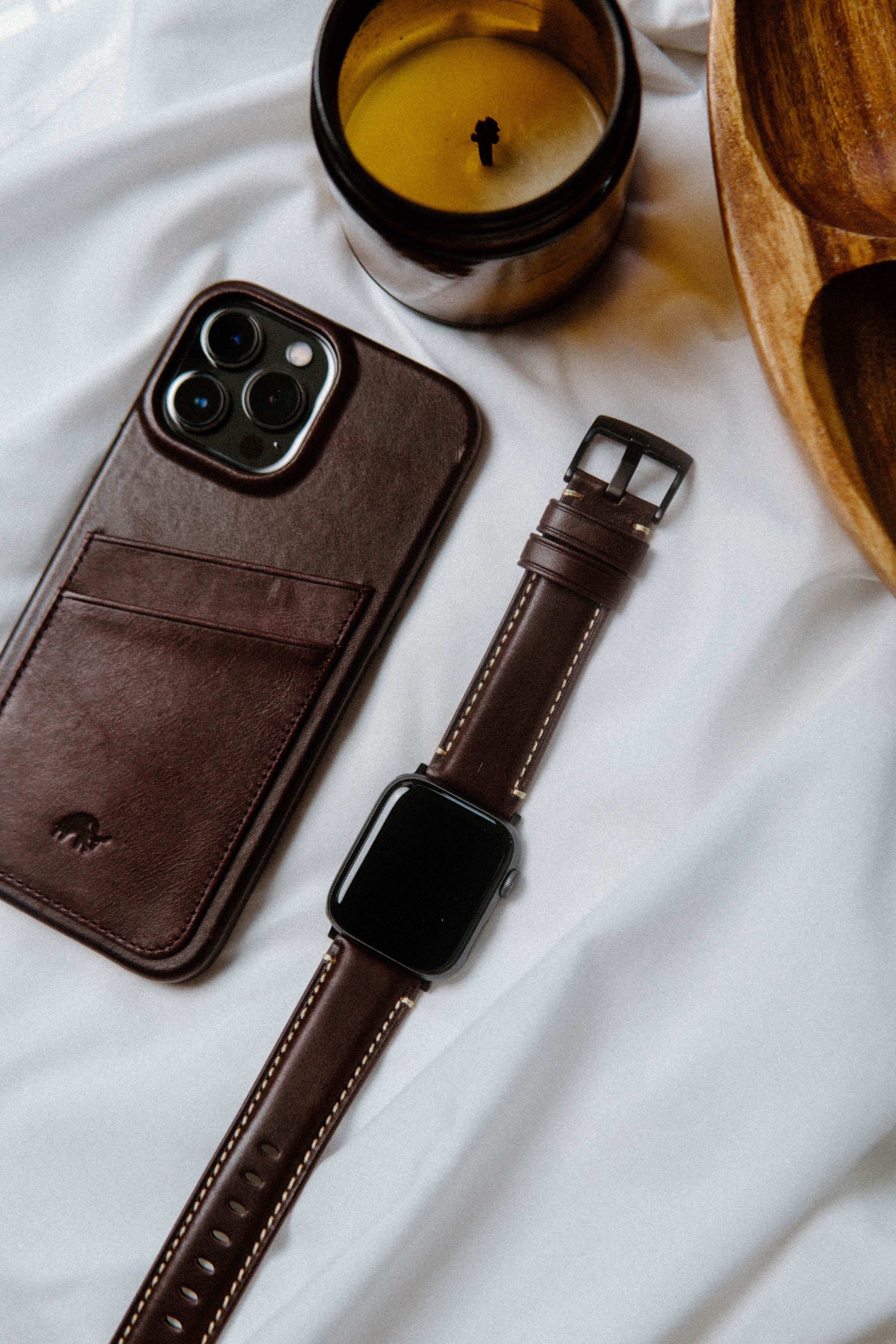 A leather Apple Watch strap next to a phone case and a candle on a white surface.