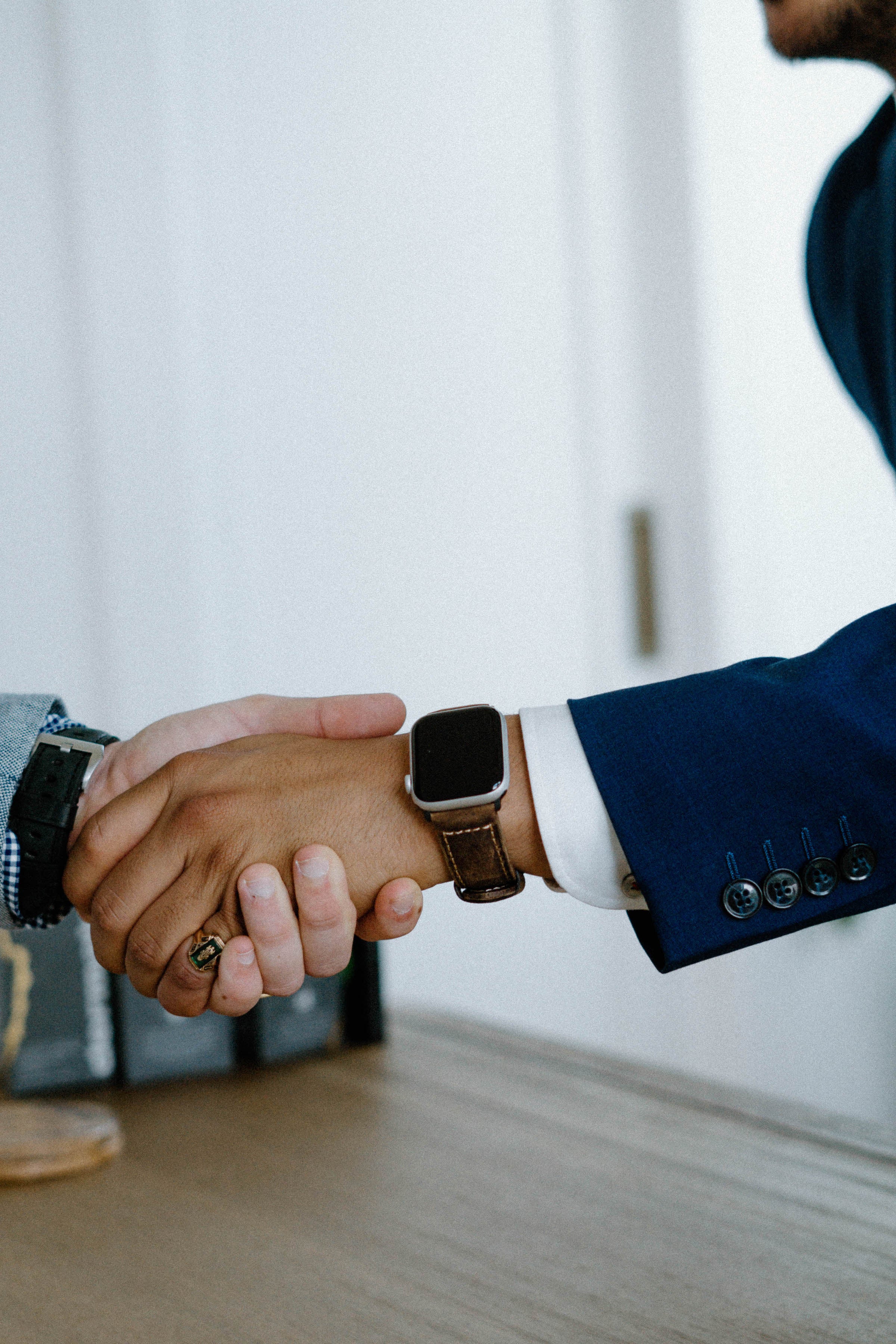 Two people shaking hands across a desk; visible blue suit sleeve and leather-banded smartwatch on one wrist.