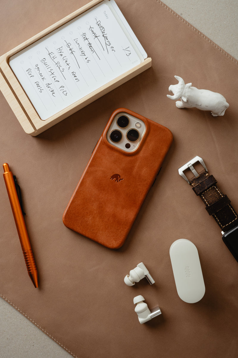 Everyday carry flatlay: brown leather phone, notebook showing 'Today' and '1/3', orange pen, watch, white earbuds, ceramic elephant.