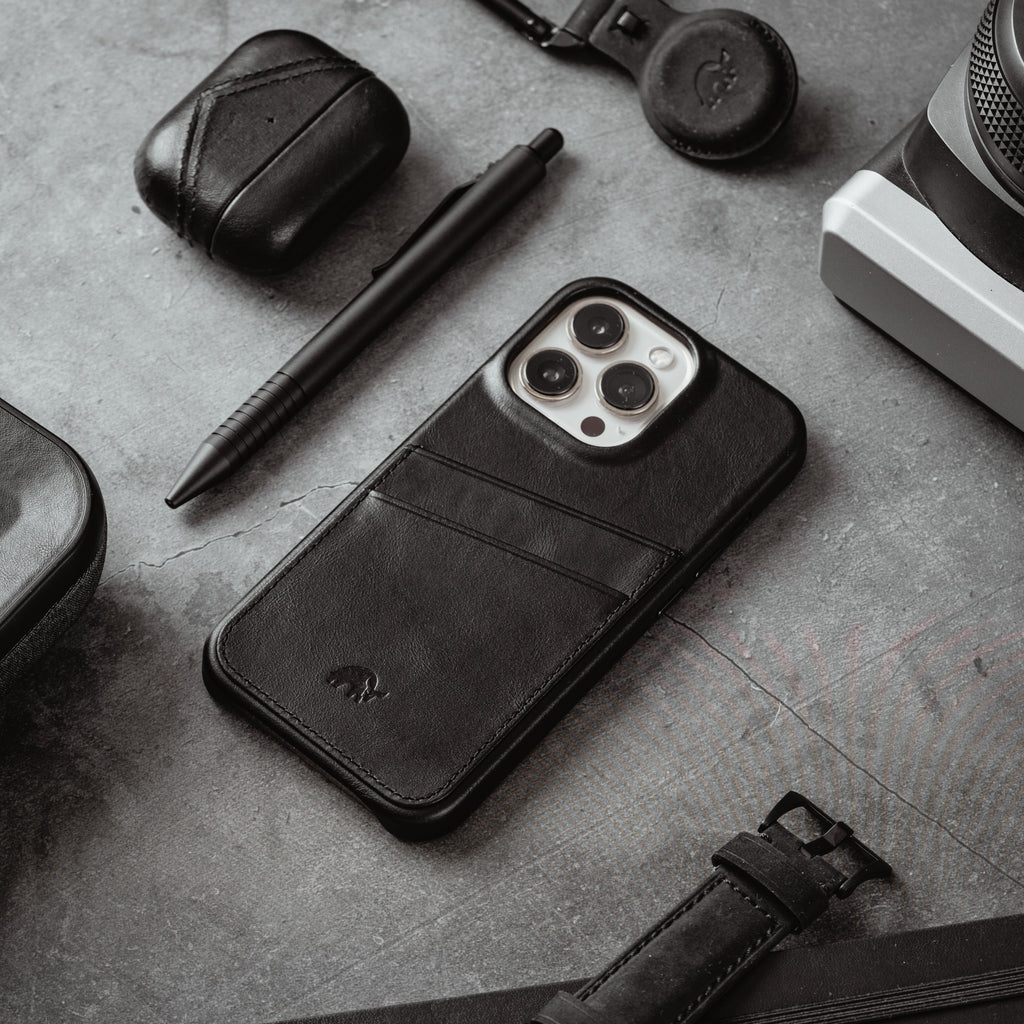 Black leather phone case with two card slots, styled with pen, earbuds case, camera and watch strap on textured surface.