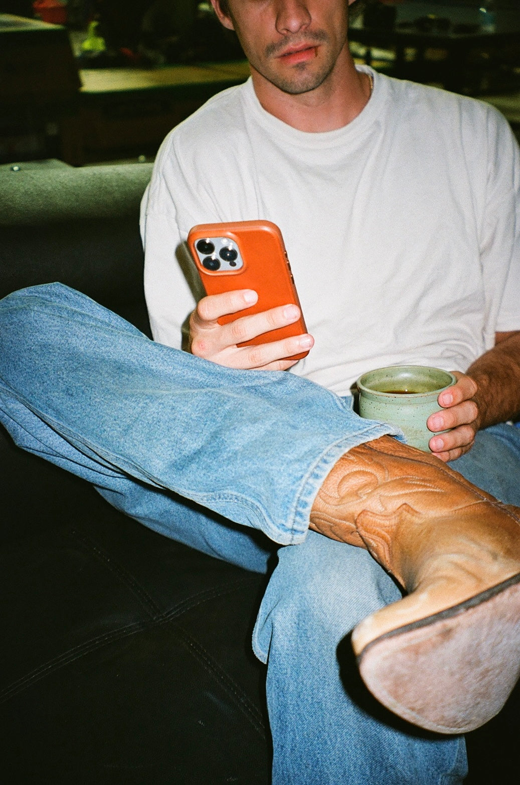 Seated man cropped at the nose, holding an orange phone and green ceramic mug; wearing a white T-shirt, blue jeans and tan cowboy boots.