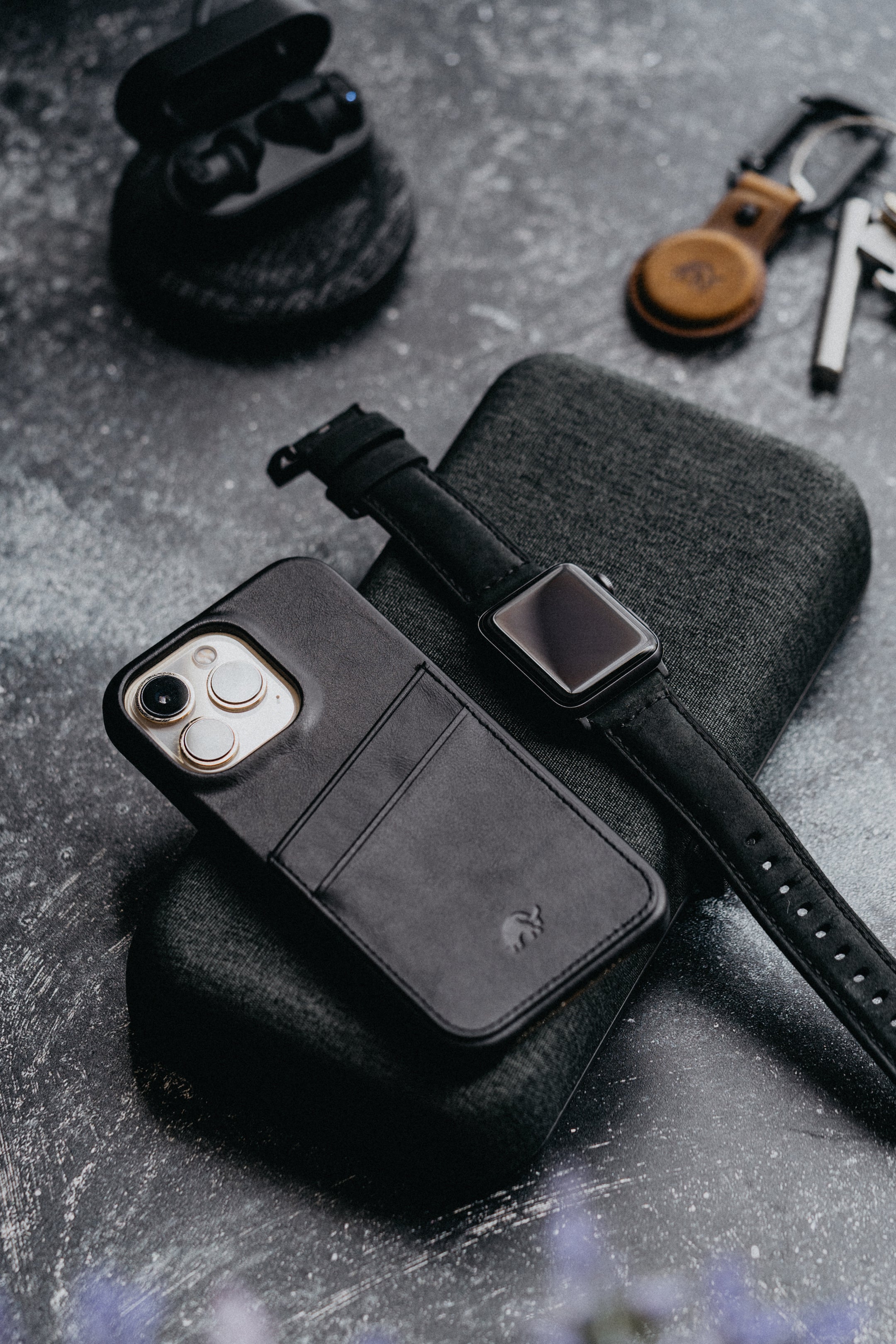 A black minimalist portfolio case with card slots, placed on a textured surface alongside a smartwatch and earbuds.