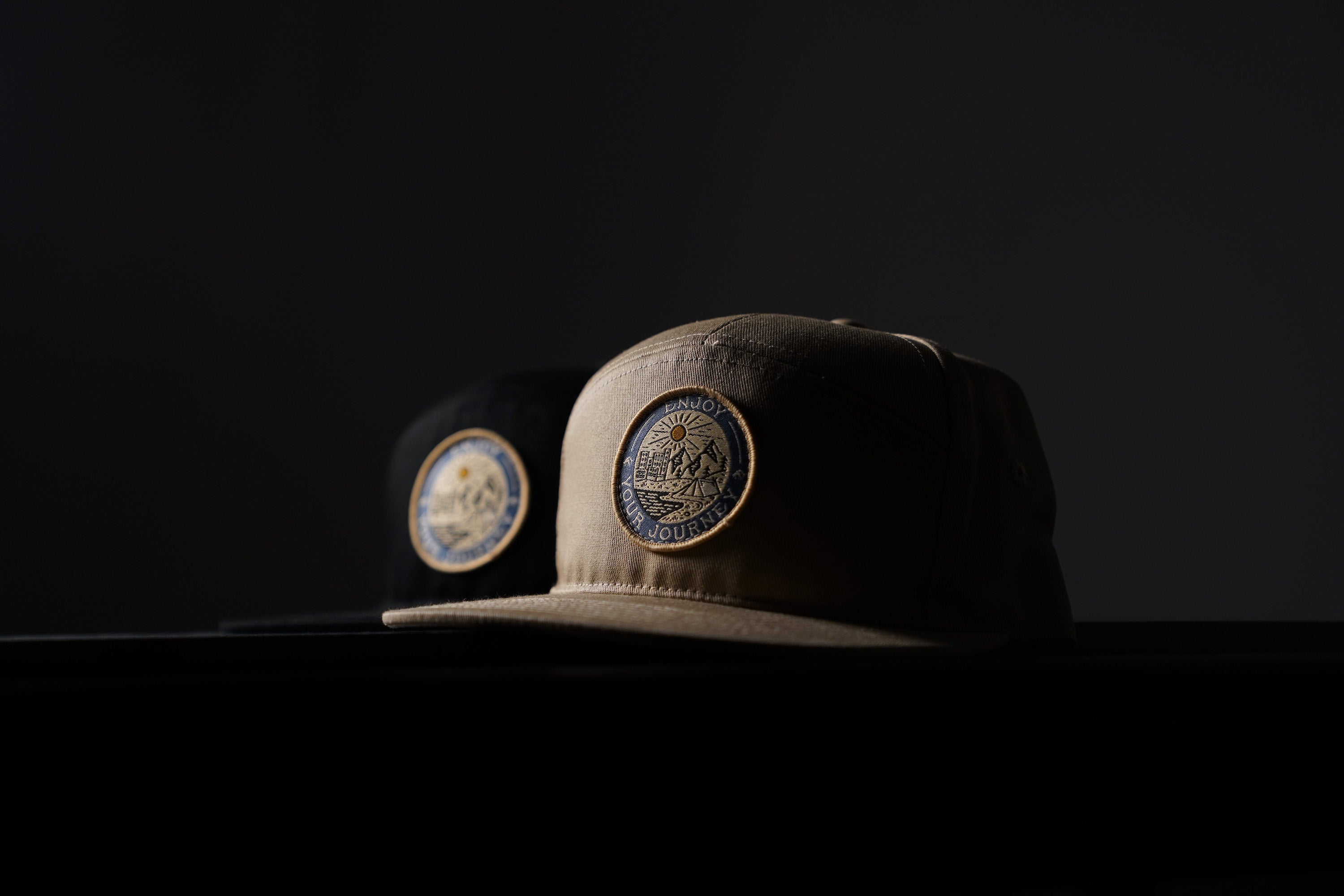 Tan cap in front and black cap behind, each with a round patch reading "ENJOY YOUR JOURNEY", on a dark background.