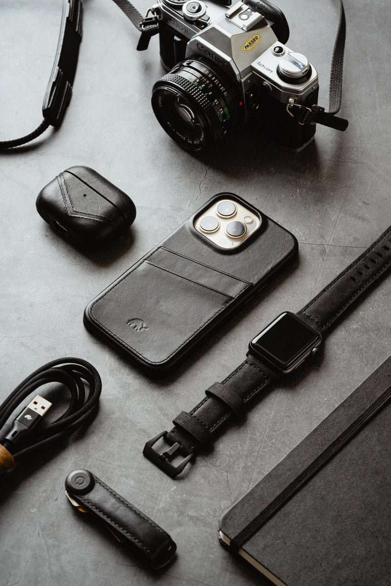 A flat lay of a camera, leather phone case, AirPods, watch, charging cable, and notebook.